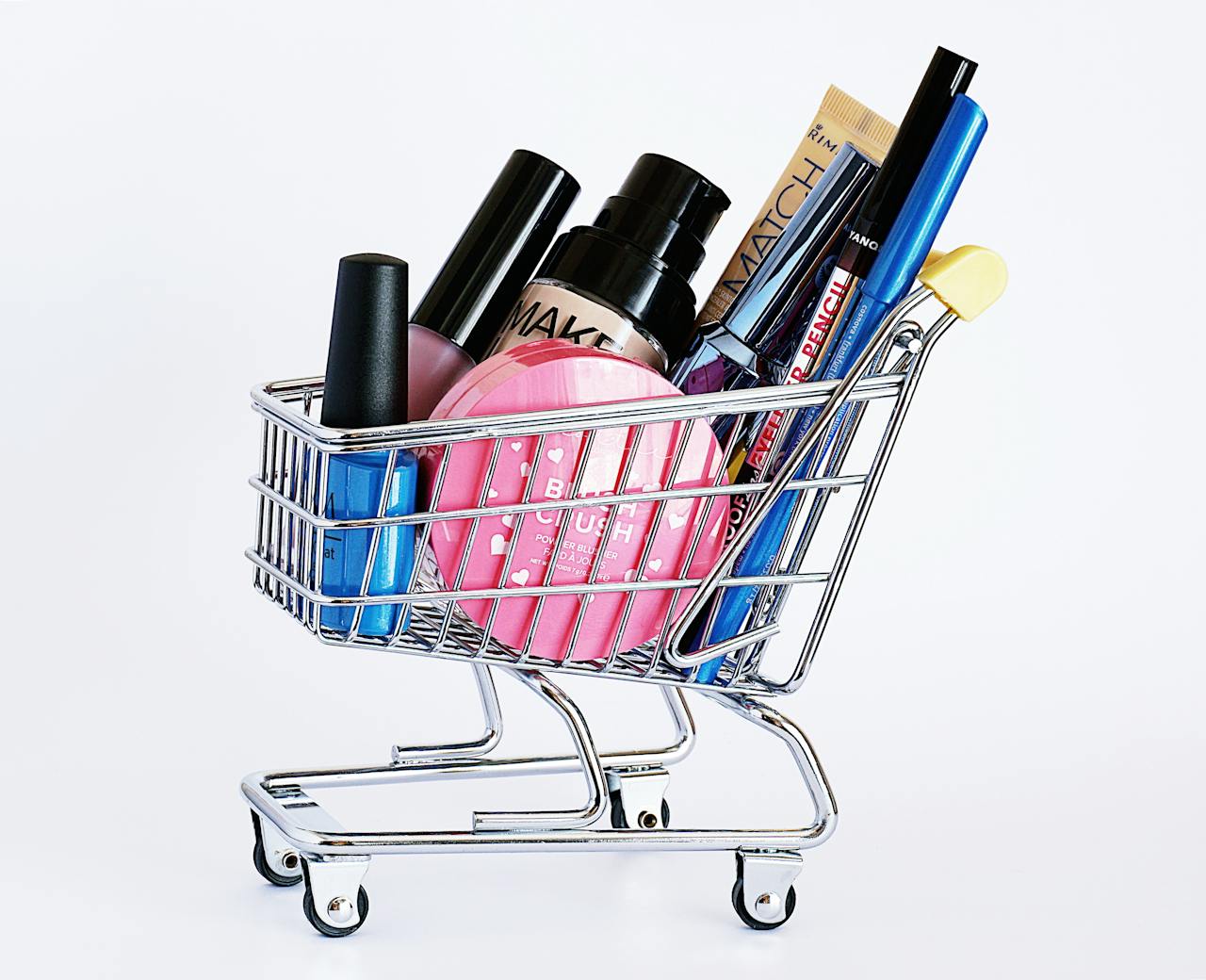 Miniature shopping cart filled with a variety of cosmetics on a white background.